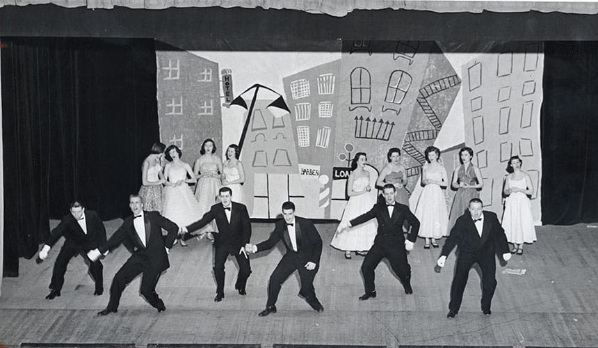 On a stage with a set backdrop of a city street, six men in tuxedos dance in front of nine women standing in formal gowns as part of a Humorology production circa 1950-1959.