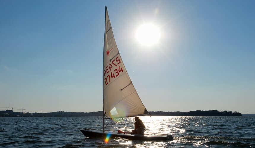 Silhouetted by summer sunlight shimmering off water, a Hoofer Sailing Club member sails a laser sailboat on Lake Mendota. (Image courtesy of University Communications, #9971.) A Hoofer Sailing Club member sailing on Lake Medota.