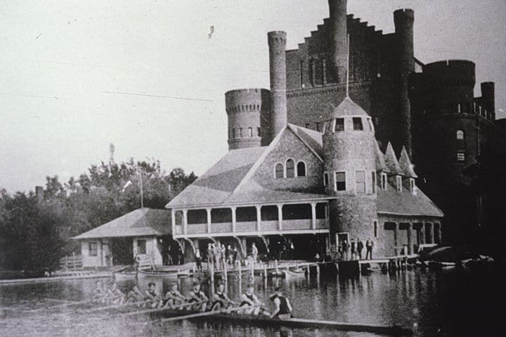 A men's crew team rows in front of the old boathouse and Armory, as spectators look on from the pier. Image couresy of UW Archives, #S11578